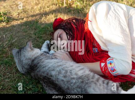 Junge Frau mit scharlachroten Dreadlocks in Nationaltracht auf dem Gras liegen und mit der Katze spielen. Außenportrait Stockfoto