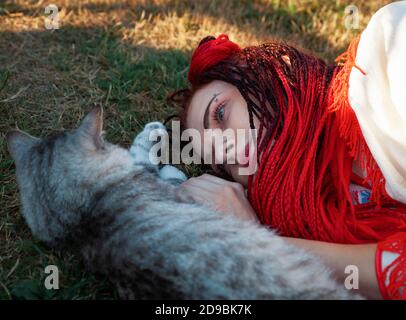 Junge Frau mit scharlachroten Dreadlocks in Nationaltracht auf dem Gras liegen und mit der Katze spielen. Außenportrait Stockfoto