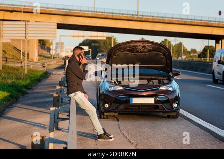 Junger Mann, der in der Nähe eines kaputten Autos steht und um Hilfe oder Hilfe ruft. Rufen Sie zum Automechaniker für Auto-Reparatur. Stockfoto