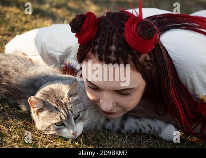 Junge Frau mit scharlachroten Dreadlocks in Nationaltracht auf dem Gras liegen und mit der Katze spielen. Außenportrait Stockfoto