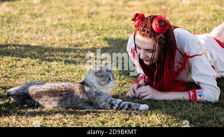 Junge Frau mit scharlachroten Dreadlocks in Nationaltracht auf dem Gras liegen und mit der Katze spielen. Außenportrait Stockfoto