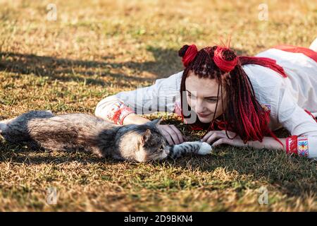 Junge Frau mit scharlachroten Dreadlocks in Nationaltracht auf dem Gras liegen und mit der Katze spielen. Außenportrait Stockfoto