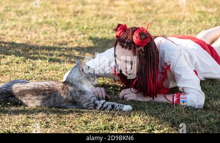 Junge Frau mit scharlachroten Dreadlocks in Nationaltracht auf dem Gras liegen und mit der Katze spielen. Außenportrait Stockfoto