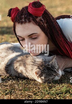 Junge Frau mit scharlachroten Dreadlocks in Nationaltracht auf dem Gras liegen und mit der Katze spielen. Außenportrait Stockfoto