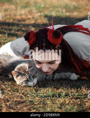 Junge Frau mit scharlachroten Dreadlocks in Nationaltracht auf dem Gras liegen und mit der Katze spielen. Außenportrait Stockfoto