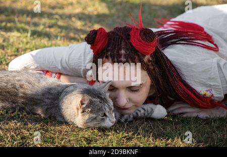 Junge Frau mit scharlachroten Dreadlocks in Nationaltracht auf dem Gras liegen und mit der Katze spielen. Außenportrait Stockfoto