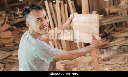Der Zimmermann lächelt und schaut auf die Kamera, während er einen hält Holzdiele, um den Stuhl wieder in der zu befestigen Werkstatt Stockfoto
