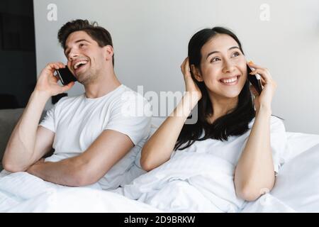 Foto eines fröhlichen jungen liebenden Paares zu Hause Im Schlafzimmer im Bett liegend, während man mit dem Handy spricht Telefone Stockfoto