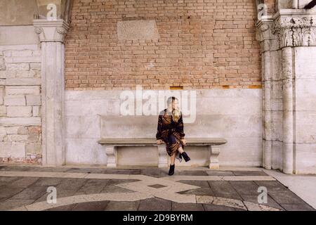 Schönes Mädchen mit einem braunen und schwarzen langen Kleid, sitzt auf einer Bank und schaut zur Seite, in Venedig, Italien Stockfoto