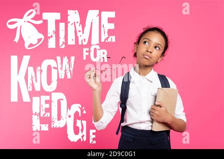 Nachdenkliche afroamerikanische Schulmädchen mit Buch und Brillen in der Nähe stehen Zeit für Wissensschrift auf Pink Stockfoto