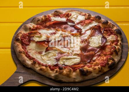 Pizza mit Speck oder Speck und Brie-Käse und Tomatensouce. Typisches italienisches Produkt auf Holzschneidebrett auf gelbem Hintergrund Stockfoto