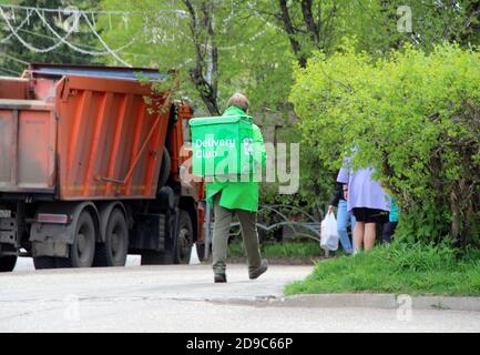 24.05.2020. Syktywkar, Russland. Der Kurier der Lebensmittelfirma 'Delivery Club' mit einer grünen Tasche hinter dem Rücken und in grünen Uniformen geht im Frühjahr die Straße der Stadt entlang vor dem Hintergrund eines LKW und Bäumen. Stockfoto