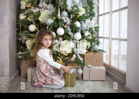 Kleines Mädchen in schönen Kleid sitzt unter Weihnachtsbaum mit Geschenkbox und Bogen. Licht von einem großen Fenster, dunklen Raum, Wohnkomfort, Weihnachten, m Stockfoto