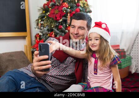 Papa und Tochter in der Nähe des Weihnachtsbaumes mit einem Smartphone machen ein Selfie, kommunizieren über Video-Verbindung. Weihnachtsgrüße, eine Geschenkbox, ein Mädchen in Stockfoto