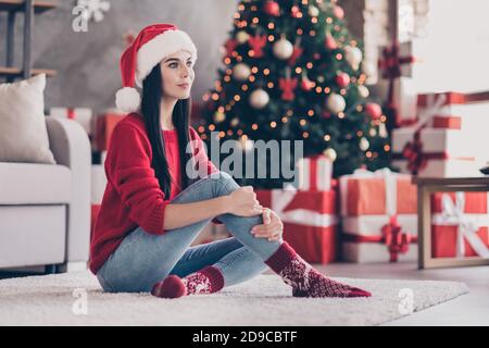 Foto von jungen charmanten Mädchen sitzen Teppich Umarmung Knie tragen santa Kopfbedeckung rote Pullover Jeans Socken im dekorierten Wohnzimmer In Innenräumen Stockfoto