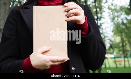 Nahaufnahme der Frau in schwarzem Wollmantel und Burgunder Pullover mit einem Buch mit leerem Einband im Park. Freier Platz für Ihr Lesekonzept Stockfoto