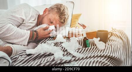 Ungesunde und müde Mann niesen oder wipping notty Nase Mann Liegen in einem gemütlichen Hausbett neben viel Gebrauchte Papiertaschentücher und Medikamente auf der Decke Stockfoto