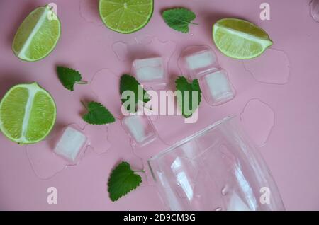 Zutaten für Mojito auf rosa Hintergrund. Leeres Glas, Stroh, Minze, Limette, Eiswürfel. Limette und Minze fallen in einen Cocktail spritzt Stockfoto