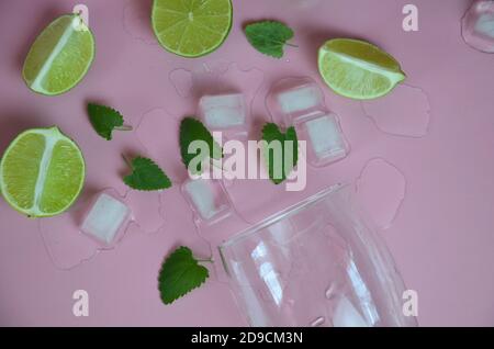 Zutaten für Mojito auf rosa Hintergrund. Leeres Glas, Stroh, Minze, Limette, Eiswürfel. Limette und Minze fallen in einen Cocktail spritzt Stockfoto