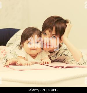 Glückliche junge Mutter und Baby Mädchen das Lesen eines Buches auf dem Bett Stockfoto