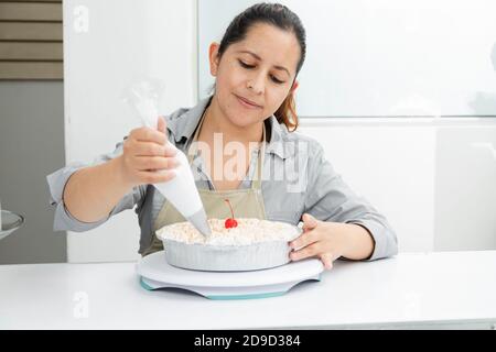 Hispanische Frau in ihrer Patisserie dekorieren einen Kuchen - Frau Putting Schlagsahne auf frisch gebackenen Kuchen - unternehmungslustige Frau Stockfoto