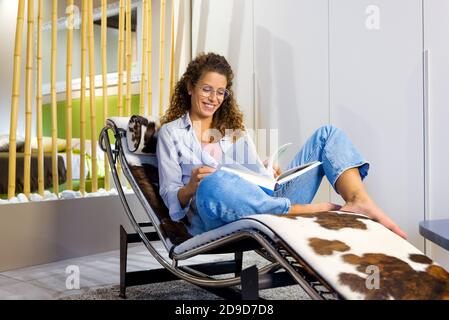 Glücklich zufrieden junge Frau trägt Brille Entspannung barfuß auf einem Stilvoller Sessel, der mit einem Lächeln ein Buch liest Stockfoto