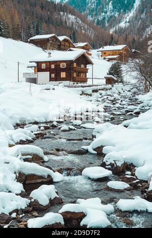 Eis- und schneebedeckter Bach an einem kalten Wintertag vor traditionellen Holzhütten des kleinen Bergdorfes St. Gertraud im Ultental, Südtirol/ Stockfoto