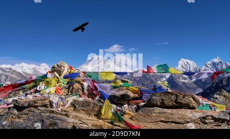 Spektakulärer Panoramablick auf das Mount Everest-Massiv vom Renjo La Pass, Himalaya, Nepal mit buddhistischen Gebetsfahnen, die im Wind fliegen und einem schwarzen Vogel. Stockfoto