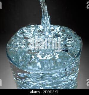 Wasser läuft mit Blasen in ein Glas, das gegen überläuft Ein dunkler Hintergrund Stockfoto