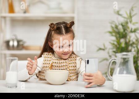 Nettes kleines Mädchen Frühstück essen: Müsli mit der Milch und suchen Cartoon in Smartphone in der Küche. Gesundes Frühstück Stockfoto