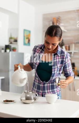 Nahaufnahme einer jungen Frau, die heißes Wasser über Kräuter-, Teeblätter hinzufügt. Lady, Lifestyle, Getränk, Zubereitung, Kräuter, Teekanne, Morgen, aromatisch, Tasse, trinken. Stockfoto