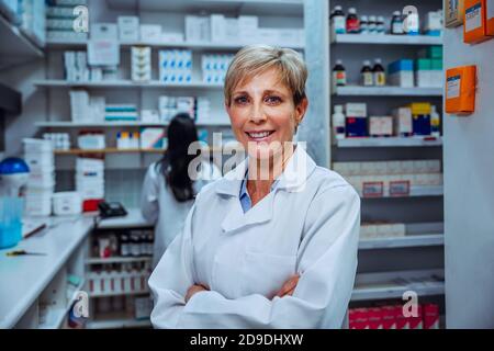 Lächelnde Apothekerin, die mit den Kreuzarmen in der Droge der Apotheke steht Speichern Stockfoto