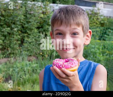 Glückliches Kind isst einen rosa Donut im Park. Ungesunde Ernährung Konzept, Naschen süße Lebensmittel. Kinder lieben süßes Essen Stockfoto