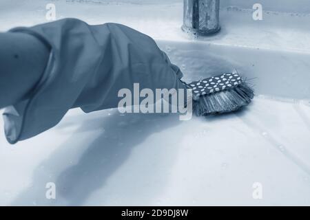 Eine Hand in einem gelben Handschuh reinigt das Badezimmer mit einer blauen Bürste auf dem Griff. Hausaufgaben, Desinfektion. Klassisches Blau Stockfoto