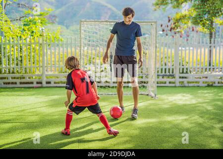 Kleiner niedlicher Junge in roter Fußballuniform und sein Trainer oder Vater spielt Fußball, Fußball auf dem Feld, im Freien. Aktives Kind macht Sport mit Stockfoto