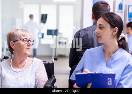 Krankenschwester, die Dokumente einreicht, während sie mit behinderten älteren Frauen im Wartebereich des Krankenhauses spricht. Patient fragt nach der Richtung an der Krankenhausaufnahme. Stockfoto