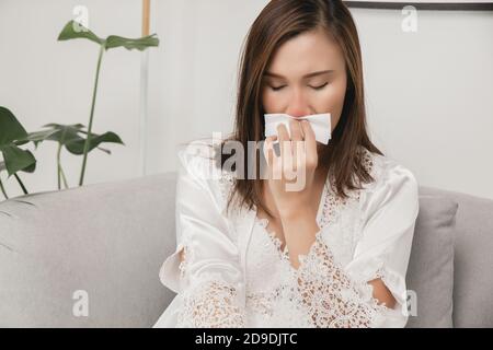 Symptome einer allergischen Rhinitis bei Frauen. Kranke Frau in weißer Nachtwäsche mit einer Erkältung, die ihre Nase zu Hause in ein Seidenpapier bläst. Allergien bei kaltem Wetter Stockfoto