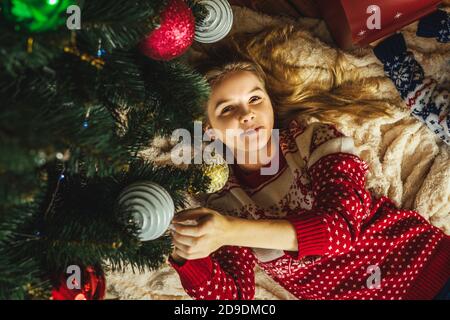 Die Frau im hässlichen weihnachtspullover liegt unter dem Weihnachtsbaum und lächelt. Neujahrskonzept. Geschmückter Weihnachtsbaum mit Kugeln. Stockfoto