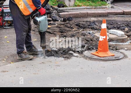 Ein Straßenarbeiter, gekleidet in reflektierende Kleidung, nutzt einen elektrischen Presshammer, um alten Asphalt von der Straße zu reißen und verwendet einen Benzingenerator, Kopierer Platz. Stockfoto