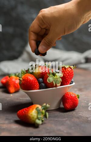 Voll von Keramikschale mit Heidelbeeren und Erdbeeren auf dem Steintisch. Und eine Hand, die Stücke von Heidelbeeren aus der Luft Stockfoto