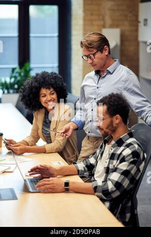 Drei multirassische Kollegen arbeiten im modernen Büro zusammen, schauen sich den Laptop-Bildschirm an und diskutieren etwas. Geschäftsleute und Teamwork-Konzept Stockfoto