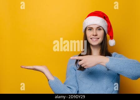 Positive junge Frau hält die Handfläche angehoben, Punkte mit Zeigefinger gegen gelbe Wand für Werbung, in Weihnachten roten Hut und blauen Pullover gekleidet Stockfoto