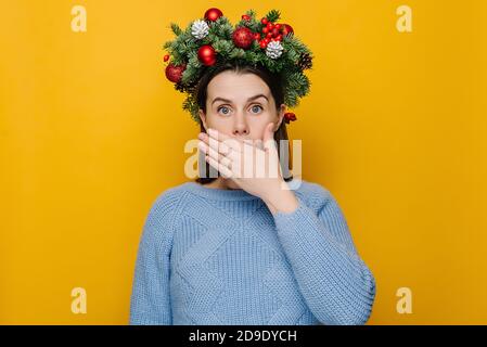 Schockiert überrascht junge Frau in traditionellen Weihnachtskranz bedeckt Mund mit Händen, tragen gemütliche blaue Pullover, isoliert auf gelbem Hintergrund Stockfoto