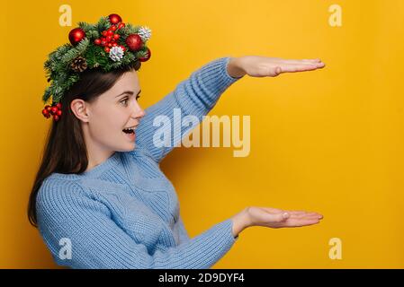 Schockiert junge Frau in Weihnachtskranz zeigt etwas großes von großer Größe Blick auf die Hände, erstaunt Mädchen mit offenem Mund überrascht mit riesigen Rabatt Stockfoto