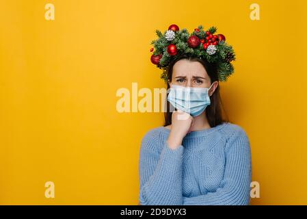 Unglückliche junge Frau trägt medizinische Gesichtsmaske, sieht traurig aus, zuckt mit gekreuzten Armen, trägt Fichtenurlaubskranz um den Kopf und Winterpullover Stockfoto