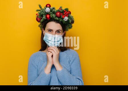 Porträt der jungen Frau in medizinischen Gesichtsmaske, unglücklich auf Kamera, trägt Fichte Urlaub Kranz um den Kopf und Winter Pullover Stockfoto