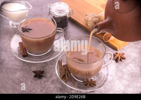 Der Prozess des Gießen von indischem Tee mit Milch und Gewürzen aus einer Keramik Teekanne in eine Tasse auf einem grauen Hintergrund. Horizontale Ausrichtung. Stockfoto