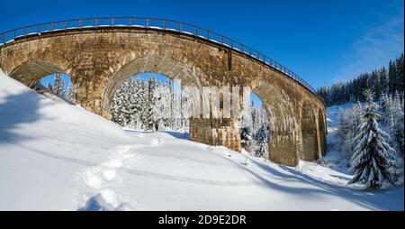 Steinviadukt (Bogenbrücke) auf der Eisenbahn durch schneebedeckten Tannenwald. Schnee driftet auf dem Weg und Raureif auf Bäumen und elektrischen Leitungen. Stockfoto