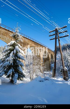 Steinviadukt (Bogenbrücke) auf der Eisenbahn durch schneebedeckten Tannenwald. Schnee driftet auf dem Weg und Raureif auf Bäumen und elektrischen Leitungen. Stockfoto