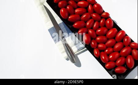 Kleine rote Kirschtomaten auf einem schwarzen rechteckigen Teller. Neben einem Cajon Handtuch und einem Stahlmesser. Platz für Text auf weißem Hintergrund kopieren. Stockfoto